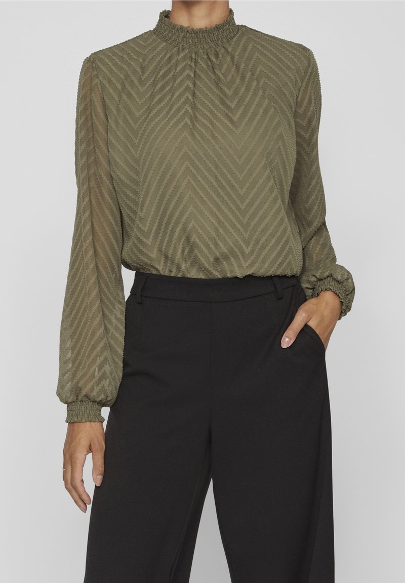 Blouse vert olive avec un motif en chevrons, un col smocké haut et de longues manches avec des poignets élastiques. Associée à un pantalon noir.