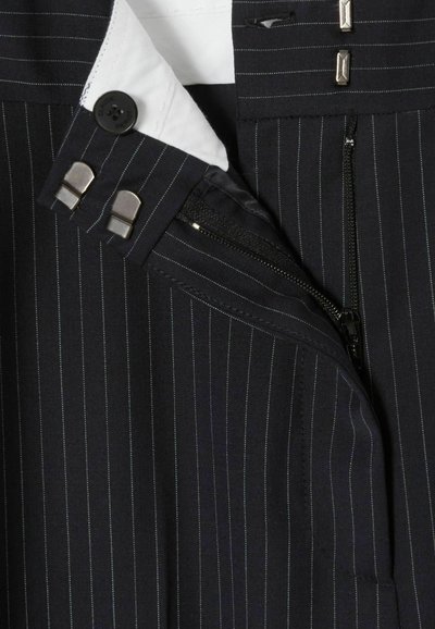 Tessuto a righe sottili blu navy con una tasca con zip, chiusura con bottoni e fodera del colletto bianca. Il design è confezionato con dettagli in metallo.