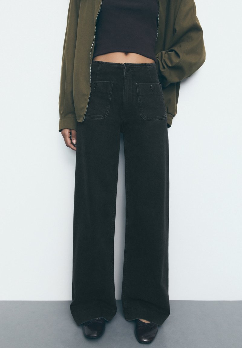 Personne portant un pantalon noir à jambes larges avec poches avant, un crop top noir, une veste zippée vert olive et des chaussures noires, debout contre un mur blanc.