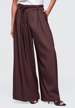 Pantalon marron à jambes larges avec plis, ceinture tressée, poches latérales, porté avec des talons ouverts noirs et un haut blanc texturé.