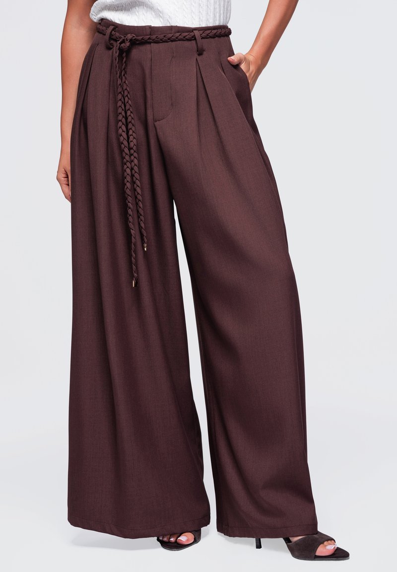 Pantalon marron à jambes larges avec plis, ceinture tressée, poches latérales, porté avec des talons ouverts noirs et un haut blanc texturé.