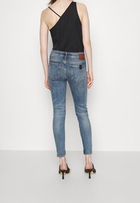 Jeans azul ajustados con una cintura de mediados de altura, bolsillos traseros y un detalle de parche de cuero marrón. Combinados con una camiseta sin mangas asimétrica negra y tacones.