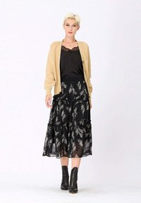 Un gilet beige sur un top en satin noir, associé à une jupe noire à volants avec des motifs de plumes argentées. Des bottines noires à la cheville complètent le look.
