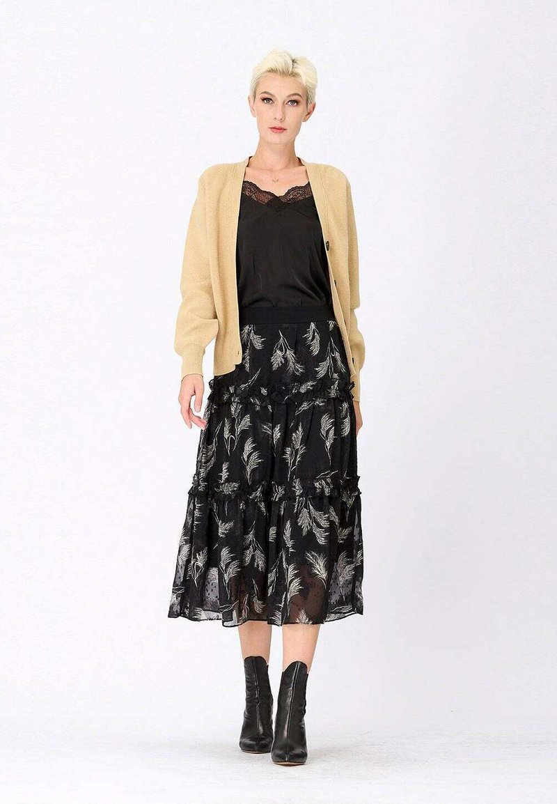 Un gilet beige sur un top en satin noir, associé à une jupe noire à volants avec des motifs de plumes argentées. Des bottines noires à la cheville complètent le look.