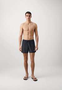 Μαύρο μαγιό σορτς με ελαστική μέση και κορδόνι, σχεδιασμένο με λογότυπο στο κάτω αριστερό μέρος. Φοριέται με μαύρες σαγιονάρες.