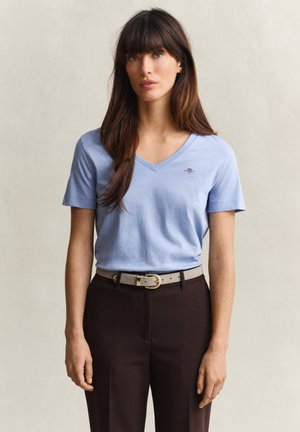 Vrouw met lang bruin haar die een lichtblauw T-shirt met V-hals en korte mouwen draagt, ingestopt in een hooggetailleerde donkere broek met een beige riem.