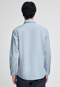 Un uomo di spalle che indossa una camicia azzurro chiaro a maniche lunghe con bottoni e pantaloni scuri, contro uno sfondo chiaro e uniforme.