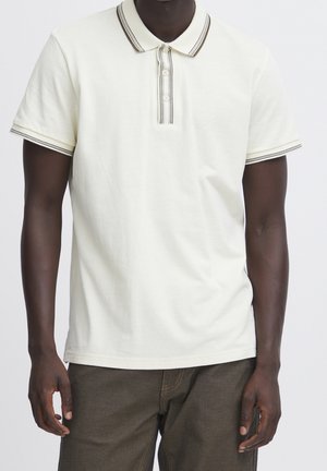 Homme portant un polo à manches courtes couleur écrue avec un col rayé et trois boutons, assorti à un pantalon marron foncé.