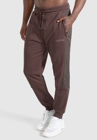 Bruin getailleerde joggingbroek met zijzakken, een trekkoord in de taille en donkergrijze zijpanelen; gestructureerde stof en eenvoudige branding zichtbaar.