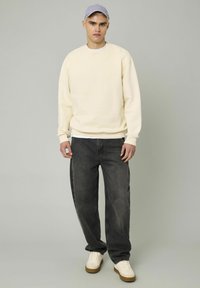 Sweatshirt crème, jeans gris et baskets blanches. Style décontracté avec ourlets côtelés. Casquette à visière plate en violet clair complète le look.