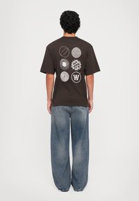 Person mit lockigem Haar, der ein dunkelbraunes T-Shirt trägt, auf dessen Rücken sechs weiße, kreisförmige abstrakte Designs abgebildet sind, sowie einer lockeren blauen Jeans.