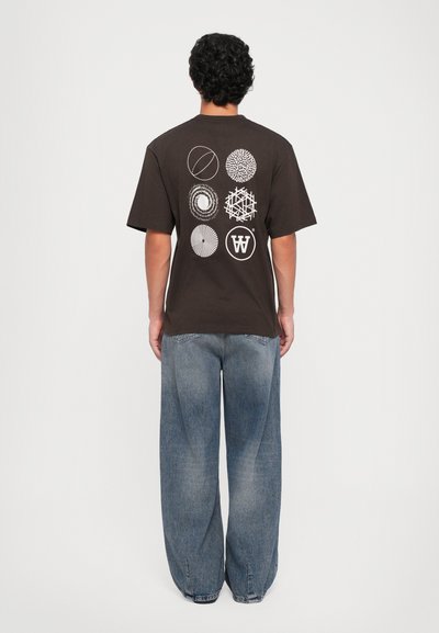 Persona con capelli ricci che indossa una maglietta marrone scuro con sei design astratti circolari bianchi sulla schiena e jeans blu larghi.