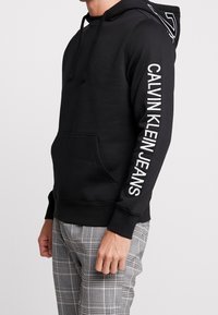 Černá mikina s kapucí s kapsou kangaroo, na pravém rukávu s bílým textem "CALVIN KLEIN JEANS" a logem na zádech. Materiál z bavlněné směsi.