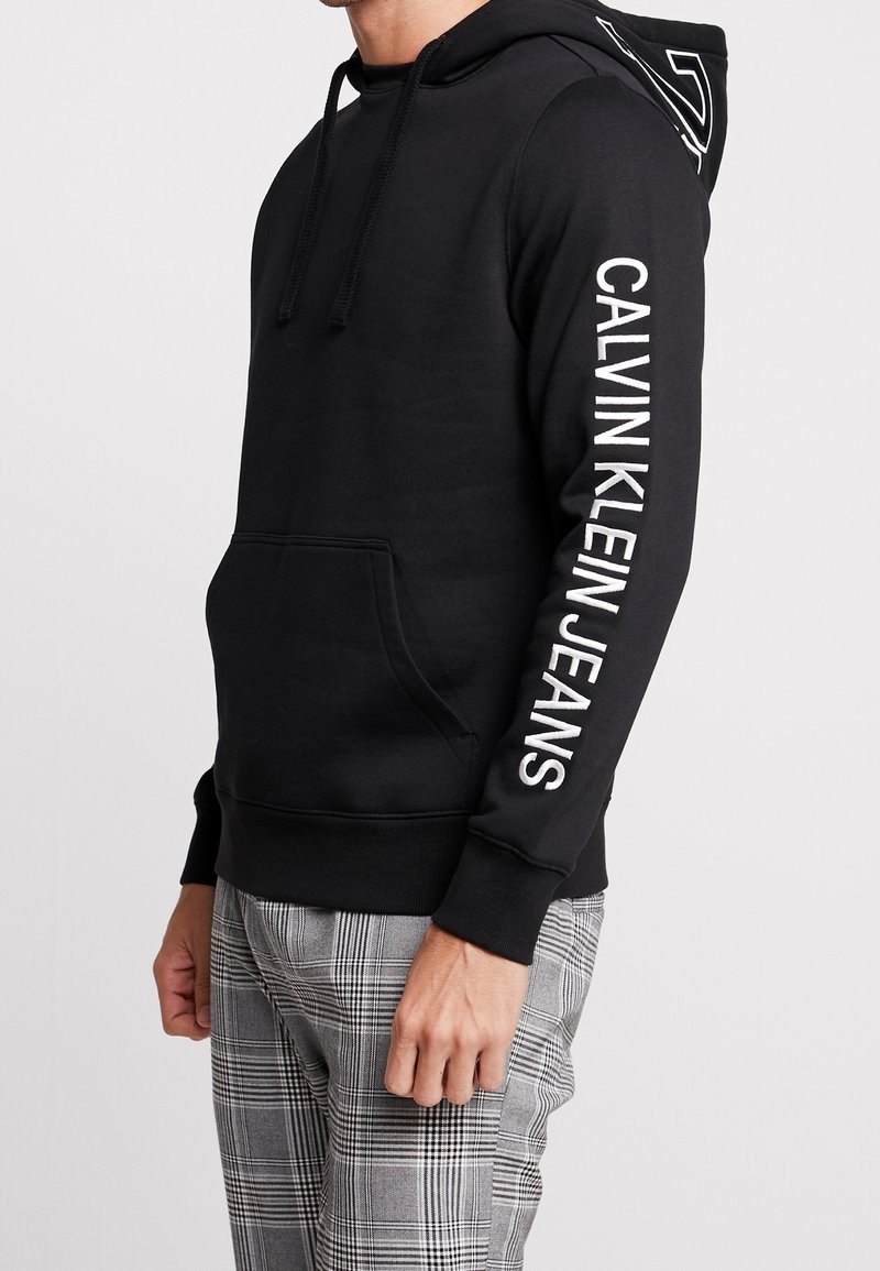 Černá mikina s kapucí s kapsou kangaroo, na pravém rukávu s bílým textem "CALVIN KLEIN JEANS" a logem na zádech. Materiál z bavlněné směsi.