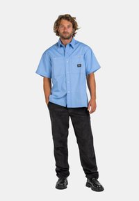 Light blue short-sleeve shirt with two chest pockets, buttons, and a logo patch. Worn with black corduroy pants and black boots.