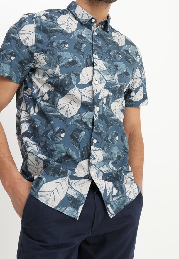 Homme portant une chemise à manches courtes à boutons avec un grand motif de feuilles bleu et blanc, associée à un pantalon bleu foncé et les mains dans les poches.