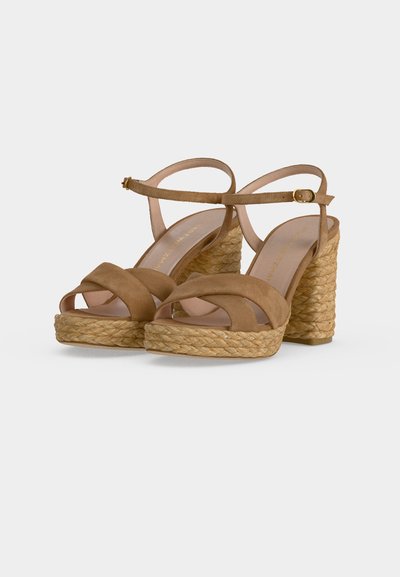Sandales compensées en daim marron avec talons compensés en corde tressée et brides de cheville avec boucles dorées sur un fond uni clair.