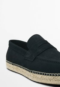 Černá semišová nazouvací bota s tkaným jute vrškem. Má plochý design, akcentované švy a široký svrchní pásek.