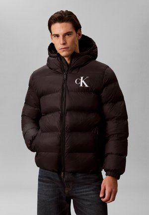 Uomo che indossa una giacca imbottita nera con cappuccio e logo "cK" sul petto, mani nelle tasche, in piedi davanti a uno sfondo grigio.