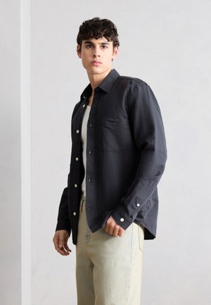 Junger Mann, trägt ein schwarzes Hemd mit Knöpfen über einem weißen T-Shirt und beige Hosen, steht vor einem schlichten hellgrauen Hintergrund.