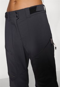 Fekete kültéri nadrág, könnyű anyagból készült, slim fit fazonú, két elülső zsebével cipzárral, és állítható derékkal.