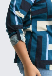 De arm van een persoon in een blauw, zwart en wit geometrisch patroon met een lange mouw, opgerold tot aan de elleboog, over een grijze t-shirt.