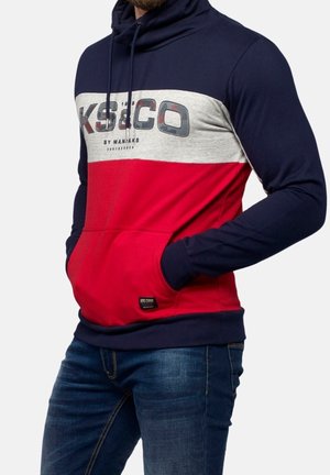 Mann trägt eine Kapuzenjacke in Marineblau, Grau und Rot mit einer Fronttasche und Logotext, kombiniert mit dunkelblauen Jeans, Hände in den Taschen.