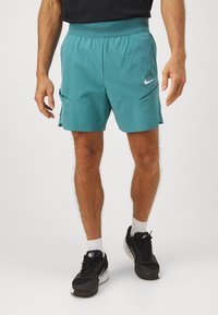 Pantalones cortos deportivos color teal, confeccionados con un tejido ligero, que presentan una cinturilla flexible, dos bolsillos laterales y un pequeño logo blanco en la parte frontal.
