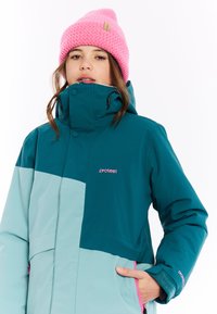 Tiefblaue und hellblaue Winterjacke mit hohem Kragen, Reißverschlusstaschen und rosa Akzenten. Gezopfter rosa Beanie mit Etikett. Strukturierter Stoff.