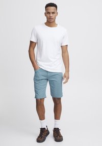 Hvitt bomulls t-skjorte, lyseblå shorts og brune sneakers med svarte detaljer. Modell som står med en hånd i lommen, nøytral bakgrunn.