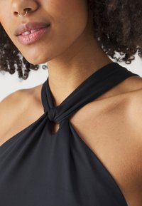Haut halter noir avec un design noué au niveau du col, présentant une découpe en forme de trou de serrure. Tissu lisse et léger qui tombe doucement sur les épaules.