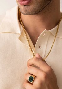 En guldkæde hænger fra en lys beige tekstureret polo shirt; en hånd holder en guld ring med en grøn sten.