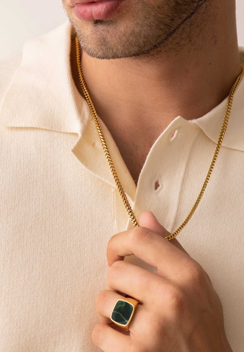 En guldkæde hænger fra en lys beige tekstureret polo shirt; en hånd holder en guld ring med en grøn sten.