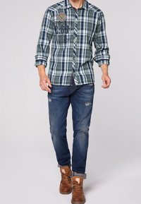 Man die wandelt in een blauwgroene geruite overhemd met knopen, versleten blauwe spijkerbroek en bruine leren laarzen tegen een effen achtergrond.