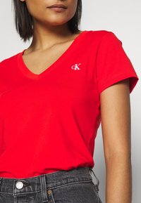 Červené bavlněné tričko s výstřihem do V a krátkými rukávy. Na levé části hrudi je bílé vyšívané logo. Kombinováno s tmavými džíny.