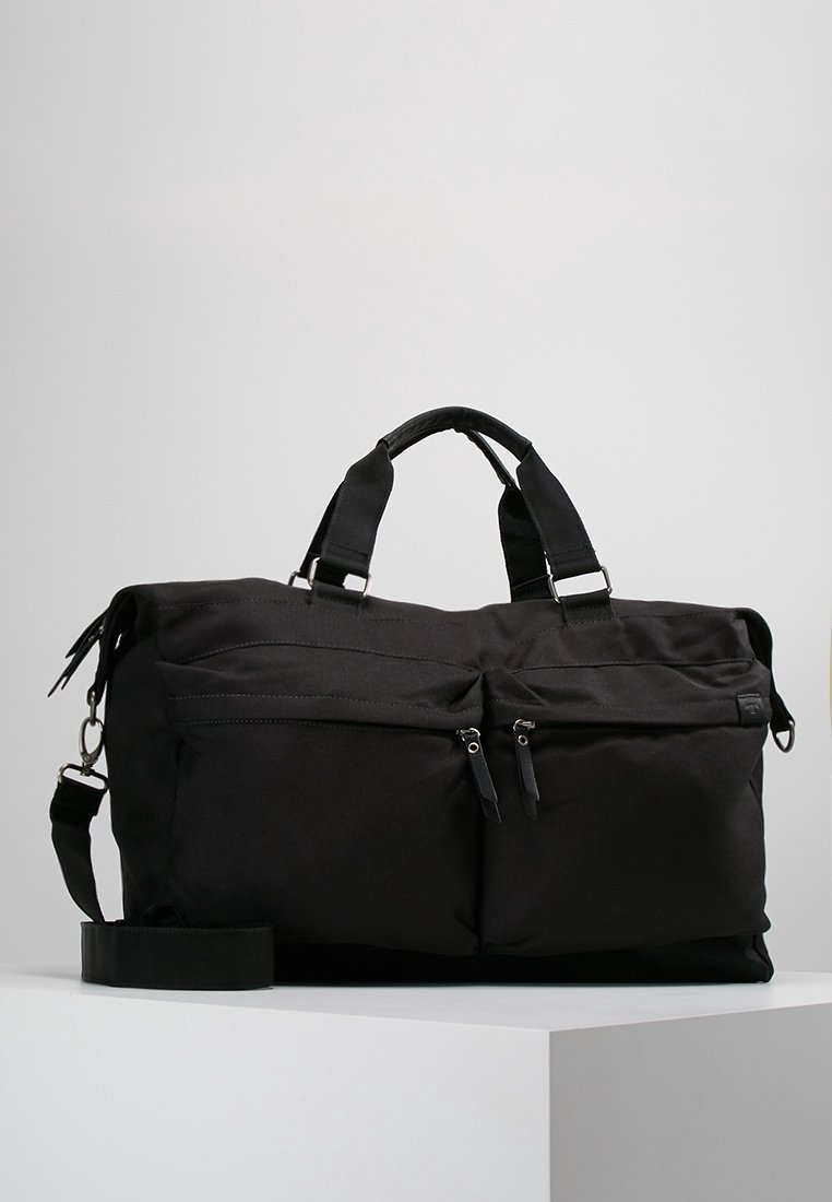 Sac de voyage souple en coque molle noire avec deux poches avant zippées, poignées robustes, bandoulière réglable et texture lisse.