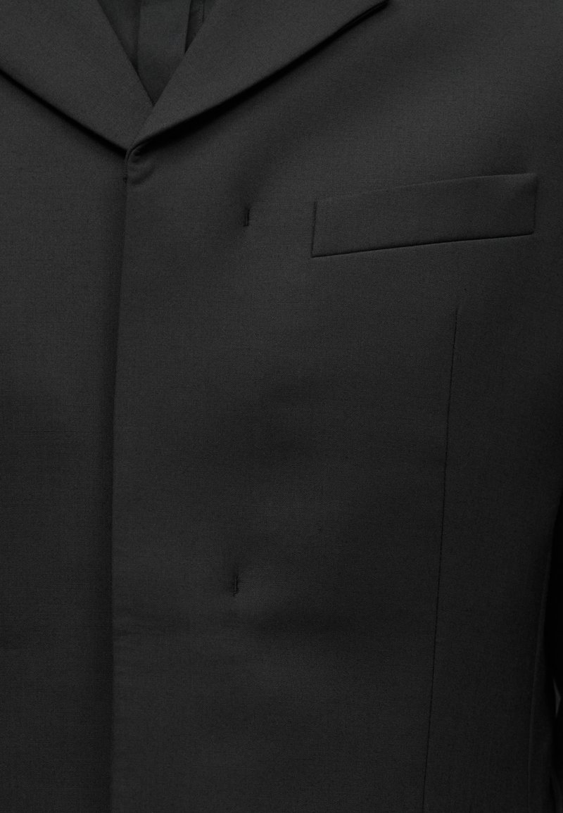 Sort jakkesætjakke med hakelapel og brystlomme med kant, knaphul syet på venstre side.