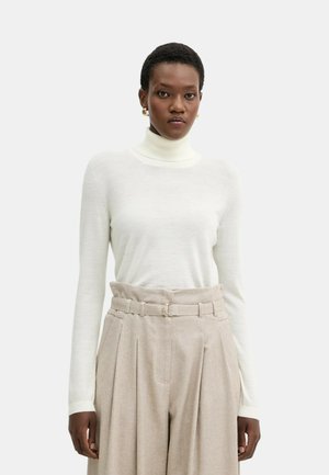 Vit långärmad polotröja i mjukt material, kombinerad med högmidjade beiga byxor med vida ben och ett bälte med rund spänne.