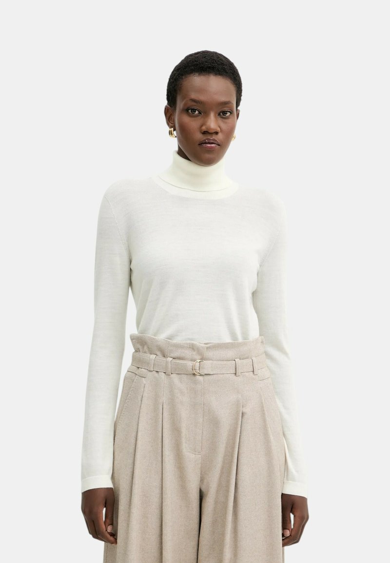 Maglia bianca a maniche lunghe con collo alto realizzata in materiale morbido, abbinata a pantaloni beige a vita alta con gambe larghe e cintura con fibbia rotonda.