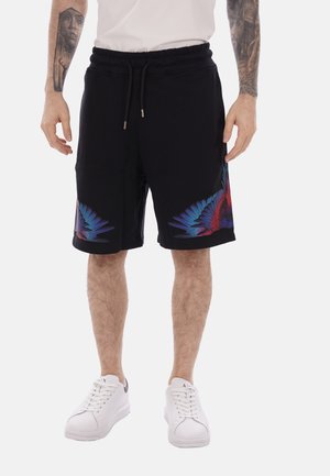Homme portant un short noir avec des motifs d'ailes bleues et rouges près des ourlets, des baskets blanches, une chemise blanche, et des tatouages visibles sur les avant-bras.