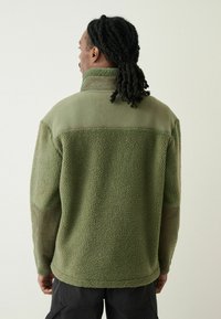 Grøn fleecejakke med høj krave, tekstureret stof og ribstrikkede ærmer. Har et delt design med bløde og glatte sektioner.