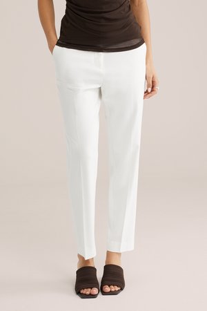 Kobieta ubrana w białe, dopasowane spodnie, ciemnobrązowy top bez rękawów oraz ciemnobrązowe, odkryte buty na obcasie typu slip-on, stojąca na jednolitym tle.