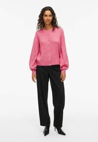 Femme portant un cardigan rose à boutons avec des manches ballon, un pantalon large noir et des talons aiguilles noirs, se tenant devant un fond blanc.