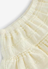 Crème-kleurige gelaagde rok met ruches aan de randen en kantafwerking, gekenmerkt door een textuurdoek en een elastische tailleband voor een verstelbare pasvorm.