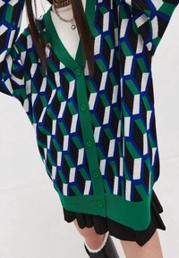 Kvinna som bär en färgglad, geometriskt mönstrad kofta med grön kant över en vit topp och svart kjol, med kedjehalsband som accessoarer.