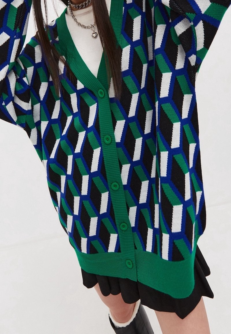 Kvinna som bär en färgglad, geometriskt mönstrad kofta med grön kant över en vit topp och svart kjol, med kedjehalsband som accessoarer.