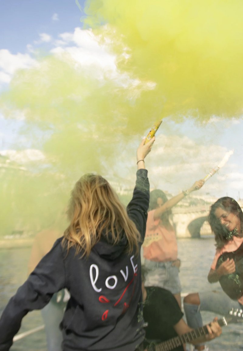 Una persona indossa una felpa nera con la scritta “love” in bianco e rosso. Fumi gialli riempiono l'aria tra un gruppo che si sta godendo una scena fluviale.