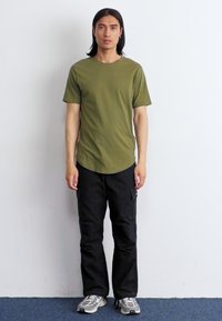 Een jonge man staat tegen een witte muur, gekleed in een olijfgroen t-shirt, zwarte broek en sneakers, met lang haar en een neutrale uitdrukking.