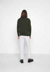 Ciemnozielony, strukturowany sweter na osobie, w połączeniu z białymi spodniami i czarnymi butami. Projekt zawiera prążkowany kołnierz i mankiety.