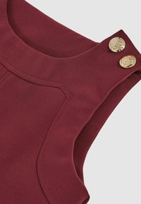 Burgundi ujjatlan felső, kerek nyakkivágással, két arany dekoratív gombbal és részletes varrással az élén.